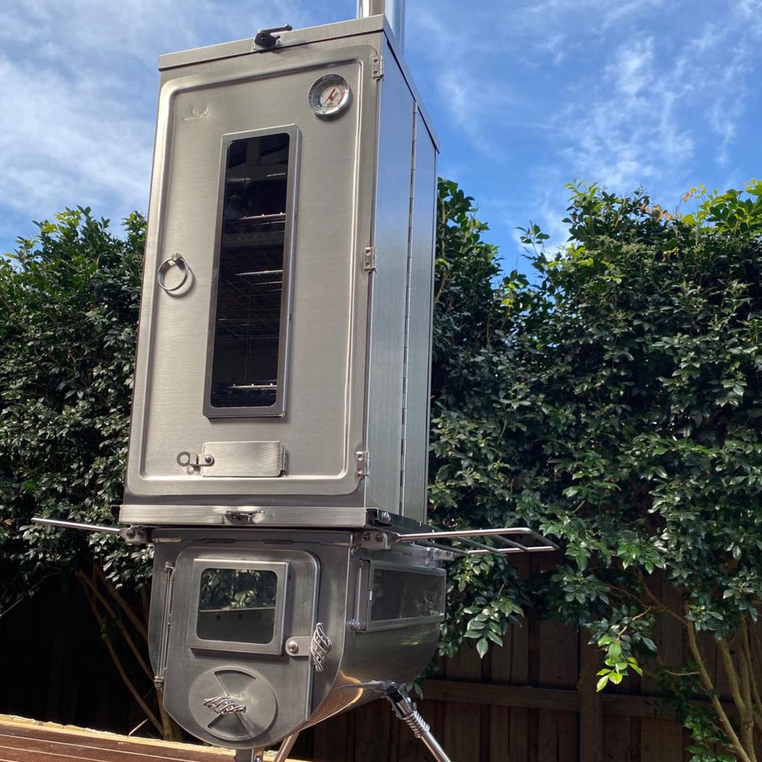 Winnerwell® Fastfold Smoker Large in outdoor use—set up in a garden/campsite, showcasing its practical application for smoking food. The large - capacity design and foldable structure make it perfect for outdoor enthusiasts hosting gatherings or preparing meals in the wild. Made of stainless steel for reliable performance.