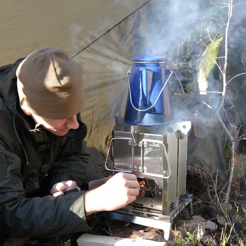 A man in a red coat, brown pants and a hat is operating the Winnerwell® Mini Multi - functional BBQ Smoker outdoors in a camping setting. The scene is set against a backdrop of a forest at twilight, with a red canoe on the right side having white letters on it. The overall color tone is cold, dominated by dark blue and gray - green, and the fire from the smoker adds a warm yellow touch. The smoker is made of durable AISI 304 stainless steel, suitable for outdoor cooking, smoking and grilling during camping