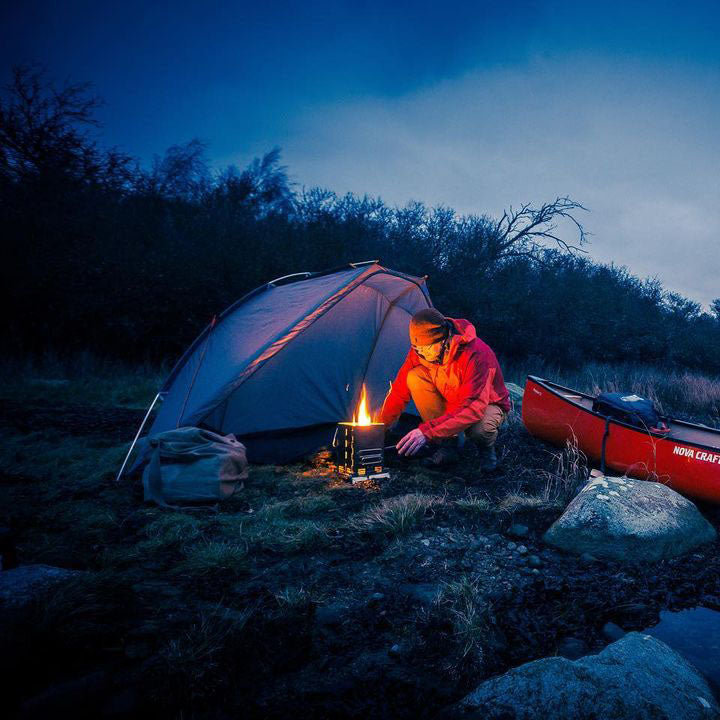 A man in a red coat, brown pants and a hat is operating the Winnerwell® Mini Multi - functional BBQ Smoker outdoors in a camping setting. The scene is set against a backdrop of a forest at twilight, with a red canoe on the right side having white letters on it. The overall color tone is cold, dominated by dark blue and gray - green, and the fire from the smoker adds a warm yellow touch. The smoker is made of durable AISI 304 stainless steel, suitable for outdoor cooking, smoking and grilling during camping