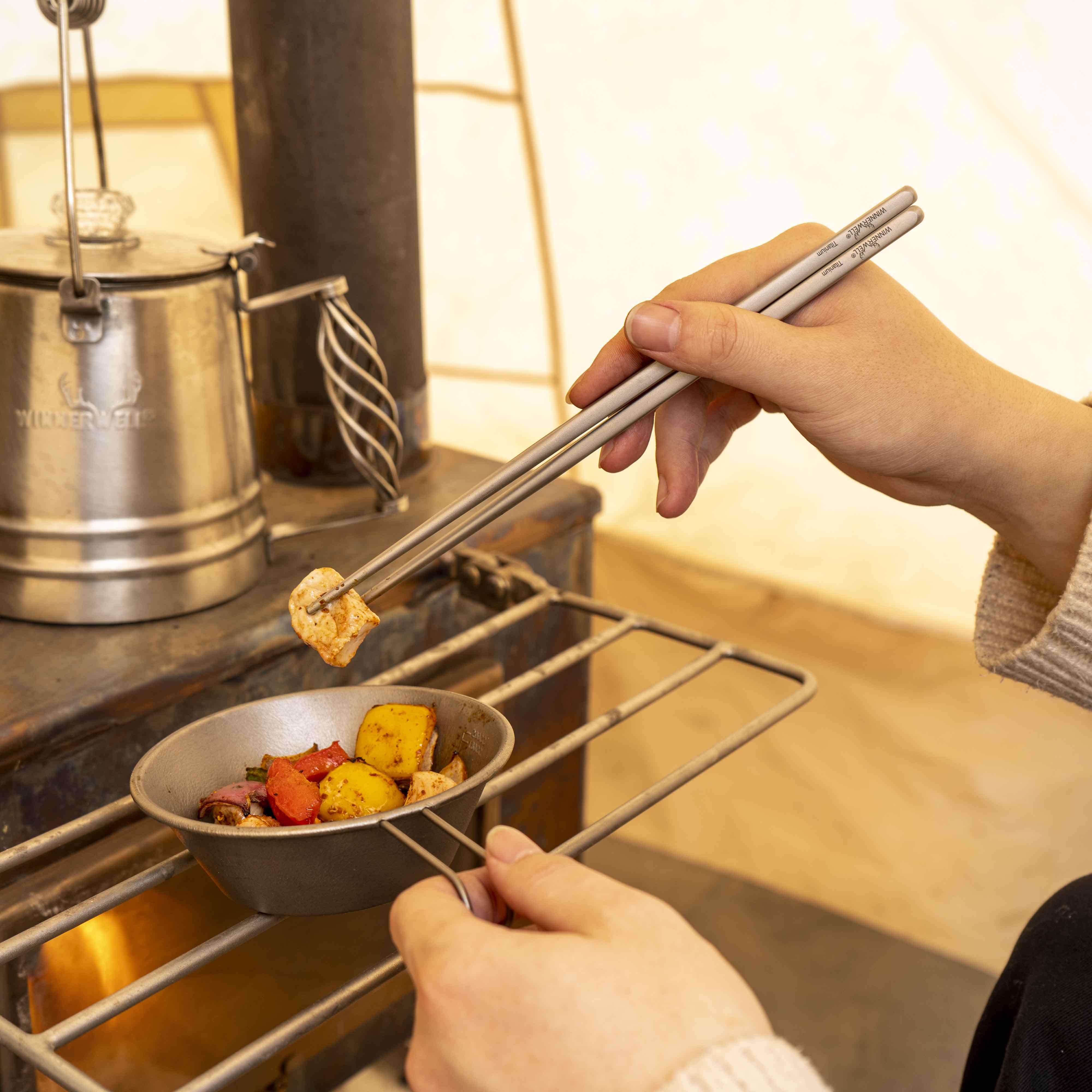 Hands using Winnerwell® square titanium chopsticks to cook food on a grill, demonstrating their practical application in outdoor cooking.