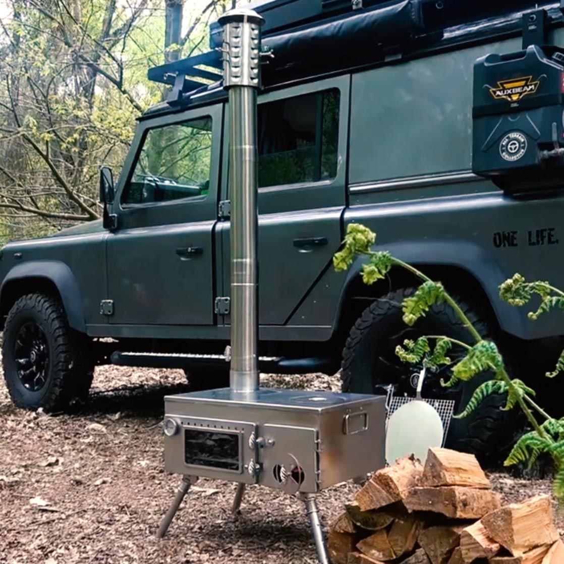 Winnerwell® Woodlander Pizza Oven Stove in use at a camping site—surrounded by nature, this stainless steel stove provides efficient, smokeless wood-burning heat for cooking pizzas, meals, or heating.